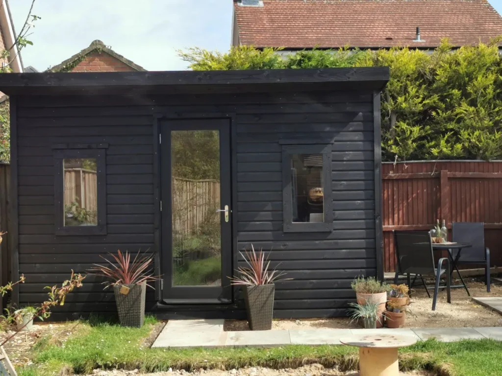 Bespoke black garden building with large front door and windows, surrounded by potted plants and outdoor seating, illustrating custom joinery and carpentry services.
