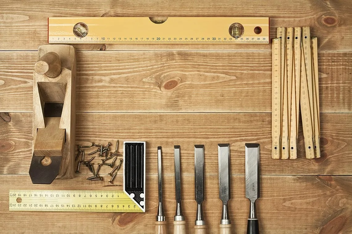 Woodworking tools arranged on a wooden surface, including a plane, measuring tape, screws, a square, and various chisels, illustrating craftsmanship and joinery for custom woodwork.