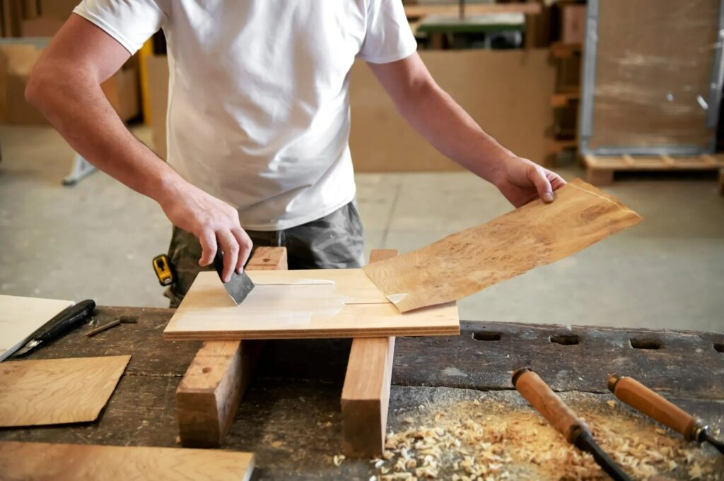 Craftsman working with wood in a workshop, using a hand tool on a wooden board, surrounded by various wood pieces and shavings, illustrating bespoke carpentry and joinery skills.