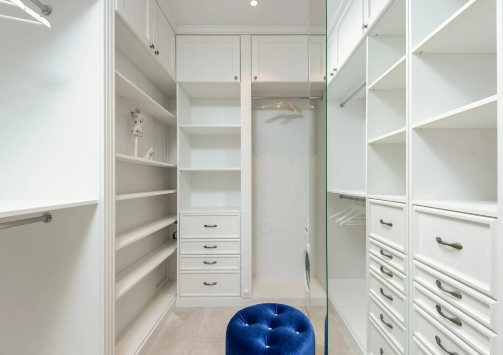 Bespoke fitted wardrobe interior with white cabinetry, shelving, and a blue upholstered stool, highlighting custom joinery and tailored storage solutions for UK homes.